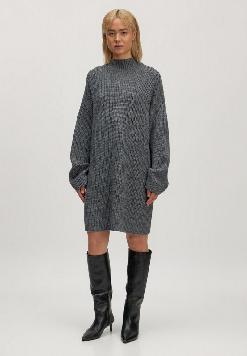 Graues Strickkleid mit langen Ärmeln, hohem Kragen und gerippter Textur, kombiniert mit knielangen schwarzen Stiefeln mit Absatz.