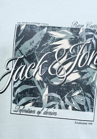 Texte "Jack & Jones" sur un motif abstrait de feuilles tropicales noires et grises avec les phrases "Définition du denim" et "Notre archive d'histoires de design."