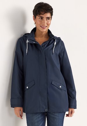 Frau trägt einen marineblauen Kapuzen-Regenmantel mit Kordelzug und aufklappbaren Fronttaschen, lächelt und blickt leicht zur Seite vor weißem Hintergrund.