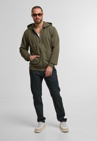 Oliwkowa bluza z kapturem na zamek błyskawiczny, z regulowanym kapturem, przednią kieszenią i miękką teksturą. Zestawiona z ciemnymi spodniami i jasnymi trampkami.