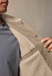 Jachetă bej căptușită cu fleece, cu un exterior moale și accente cu capse. Dispune de un buzunar în față și o cămașă gri în contrast dedesubt.