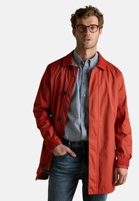 Raudona vandeniui atspari striukė su apykakle, sagomis ir laisvu siluetu, dėvima ant mėlynos ir baltos dryžuotos marškinėlės ir džinsų.