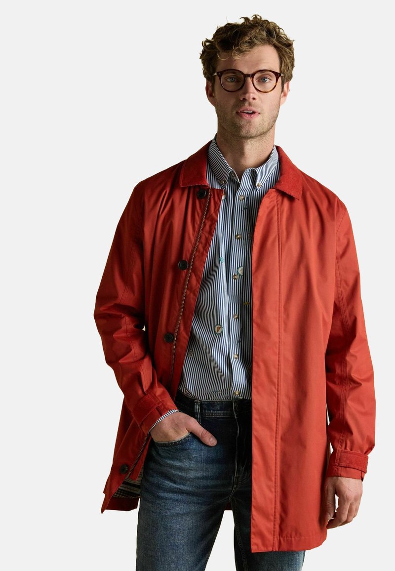 Raudona vandeniui atspari striukė su apykakle, sagomis ir laisvu siluetu, dėvima ant mėlynos ir baltos dryžuotos marškinėlės ir džinsų.