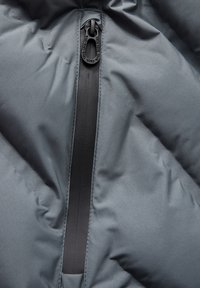 Tissu de veste en doudoune grise avec une fermeture éclair noire proéminente sur le devant, présentant une surface texturée et un design matelassé et rembourré.