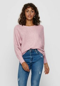 Pull en tricot rose à coupe décontractée, manches longues et poignets côtelés, associé à un jean bleu taille haute avec des détails déchirés.