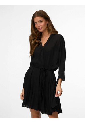 Femme aux longs cheveux bruns portant une robe noire à manches longues avec une ceinture à nouer et un décolleté en V, posant devant un fond blanc.