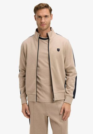 Homme portant une veste beige à fermeture éclair et un pantalon assorti avec des rayures noires et blanches sur les manches et les côtés, debout devant un fond blanc.