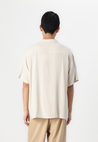 Beige short-sleeve shirt in lightweight fabric. Features a relaxed fit, back yoke seam, and a curved hem. Paired with tan trousers.