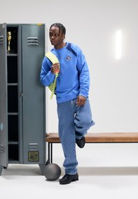 Světlé modré tričko s logem, volné džíny, černé boty a zelený batoh. Ve scéně je tmavě šedá skříňka a basketbalový míč.