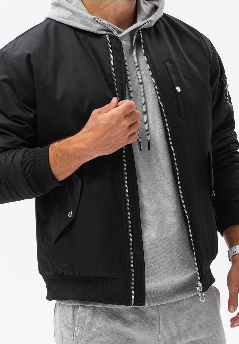 Homme portant une veste noire zippée par-dessus un sweat à capuche gris et un pantalon de survêtement gris, ajustant la fermeture éclair de la veste avec la main gauche.