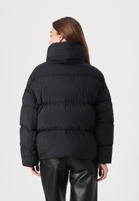 Sort pufferjakke med høj krave, quiltet design og synlige syede paneler. Båret over sorte bukser, glat tekstur med en afslappet pasform.