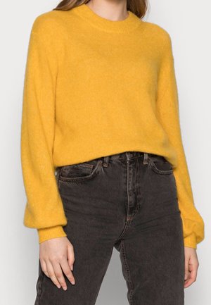 Person wearing a yellow fuzzy long-sleeve sweater tucked into high-waisted black jeans, standing with hands relaxed by sides.