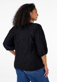 Woman with curly hair wears a textured black blouse with puff sleeves and blue jeans, standing sideways against a white background.