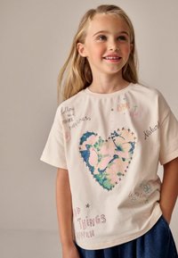 Tricou din bumbac de culoare crem, cu un design mare în formă de inimă, împodobit cu fluturi albaștri și roz, înconjurat de texte colorate și desene.