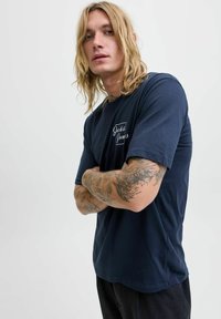 Tamsiai mėlynas medvilninis marškinėliai su trumpomis rankovėmis, su baltu logotipu krūtinėje. Audinys yra lygus, o siluetas - laisvas.