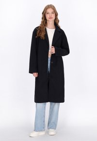 Lungo cappotto nero con colletto, chiusura con un bottone, vestibilità ampia e due tasche laterali, abbinato a jeans a gamba larga blu chiaro e scarpe bianche.