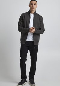Maglione grigio con zip e colletto alto, indossato sopra una T-shirt bianca. Abbinato a jeans neri e sneakers nere, in piedi su uno sfondo neutro.