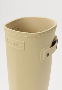 Bota de goma beige con parte superior redondeada, ribete texturizado, logo en relieve "HUNTER" y correa lateral con detalle de botón a presión. Acabado interior suave.