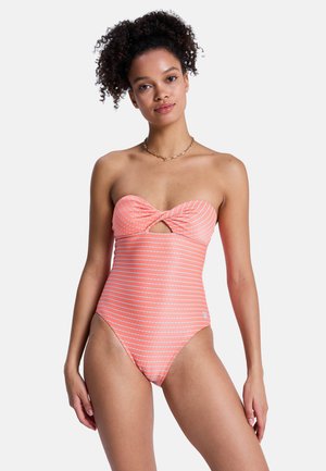 Kobieta o kręconych włosach ubrana w koralowy strój kąpielowy bez ramiączek z białymi poziomymi paskami i wycięciem z przodu, stojąca na jednolitym tle.