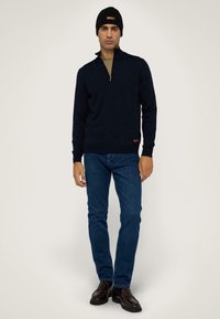 Mörkblå zip-up-tröja med långa ärmar, ihop med blå jeans och en svart stickad mössa. Inkluderar en läderdetalj på tröjans fåll.