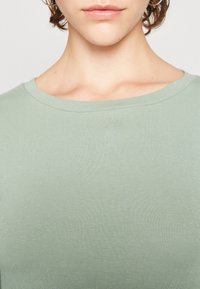 Kvinde iført en lys grøn ribbestrikket t-shirt med rund hals, synlig fra halsen til øverste bryst, med kort krøllet hår og hoops øreringe.