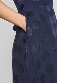 Laivastonsininen mekko, jossa on teksturoitu pinta ja suuria, tummia pilkkuja. Sivutasku näkyvissä, kangas näyttää sileältä ja siinä on hiukan kiiltoa.