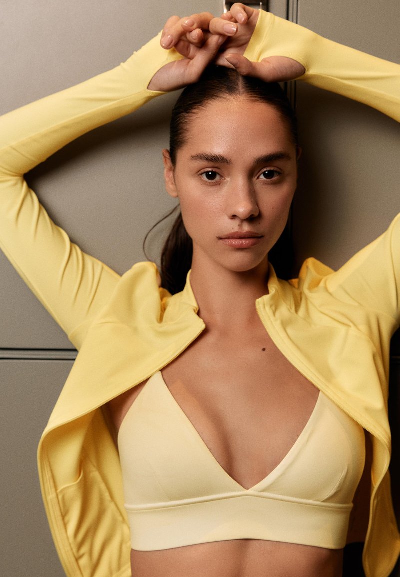 Femme en crop top jaune et veste, bras levés au-dessus de la tête sur fond beige, regardant directement la caméra avec une expression neutre.