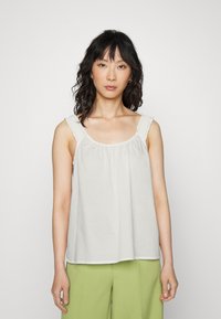 Blusa branca com alças franzidas, corte solto e decote arredondado. Usada com calças verdes de perna larga. Tecido suave e design simples.