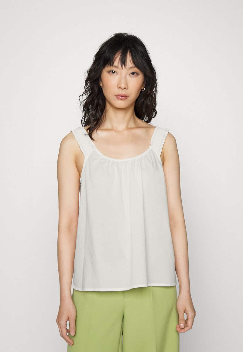 Blusa branca com alças franzidas, corte solto e decote arredondado. Usada com calças verdes de perna larga. Tecido suave e design simples.