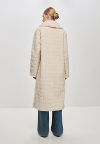 Crème-kleurige lange jas met een textuur van kabelbreiwerk, brede kraag en rechte silhouet, gecombineerd met uitlopende denim broek.