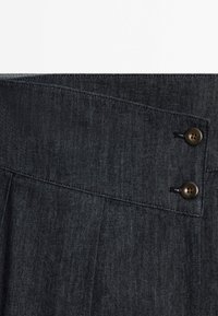 Tissu en denim foncé avec une ceinture inclinée et deux boutons ronds marron alignés verticalement sur le côté droit.