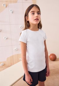 Weißes T-Shirt mit kurzen Ärmeln und rundem Ausschnitt, kombiniert mit schwarzen Shorts. Im Hintergrund ist eine Kletterwand und ein Basketball zu sehen.