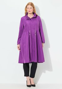 Manteau long violet, léger, avec une taille cintrée, une fermeture à boutons à l'avant et un col haut ; associé à des leggings noirs et des talons.