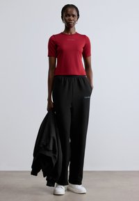 Piros, rövid ujjú póló logóval, fekete, rugalmas derékpántos melegítőnadrággal és fehér sportcipővel párosítva. A modell egy fekete kapucnis pulóvert tart a kezében.