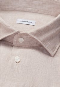 Gros plan sur le col d'une chemise en lin beige avec des boutons blancs et une étiquette "seidensticker" cousue à l'intérieur de l'encolure.