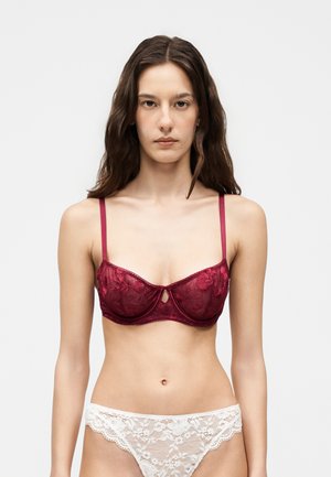 Femeie cu păr lung și castaniu poartă un sutien burgund brodat și chiloți albi din dantelă, stând în picioare în fața unui fundal simplu, deschis.