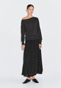 Vrouw draagt een donkere off-shoulder trui met lange mouwen, een zwarte midi-rok met patroon en zwarte slip-on puntige schoenen in een studiobeeld.