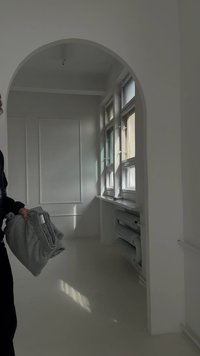 Pelēkie treniņbikses, turētas rokā, spilgti, minimālistiskā telpā ar baltām sienām, lieliem logiem un gaiša tonī grīdām.