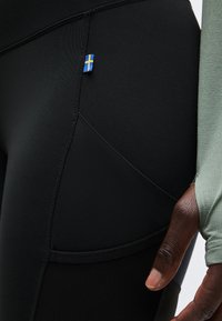 Κοντινό πλάνο μαύρων αθλητικών κολάν με μια μικρή ετικέτα της σουηδικής σημαίας και ένα χέρι μερικώς καλυμμένο από πράσινο υφασμάτινο μανίκι.