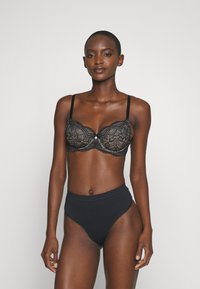 Μαύρο σουτιέν από δαντέλα με περίπλοκα σχέδια και στήριξη με μπανέλες, συνοδευόμενο από μαύρα ψηλόμεσα εσώρουχα που τονίζουν τον λείο ύφασμα και τον μινιμαλιστικό σχεδιασμό.