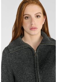 Pull en maille zippé gris anthracite avec texture côtelée, col et motif tricolore. Présente une fermeture éclair en métal et des accents.