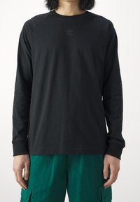 Black long-sleeve T-shirt with raglan sleeves, featuring a subtle branded logo on the chest and smooth cotton fabric. Paired with green pants.