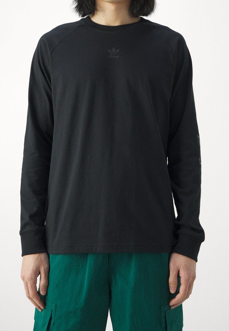 Black long-sleeve T-shirt with raglan sleeves, featuring a subtle branded logo on the chest and smooth cotton fabric. Paired with green pants.