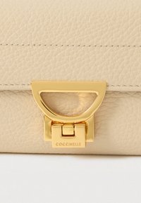 Μπεζ υφασμάτινη δερμάτινη τσάντα με χρυσό μεταλλικό κούμπωμα χαραγμένο με την ένδειξη "COCCINELLE" σε λευκό φόντο.