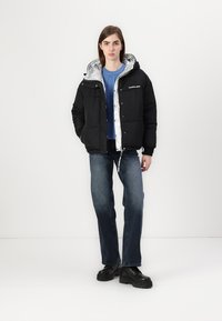 Svart puffjakke med sølvfôr, med hette og knapper foran. Brukt over en blå genser og paret med mørke jeans og svarte sko.