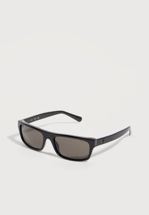 Lunettes de soleil rectangulaires noires avec des verres teintés foncés et une monture brillante, posées à plat sur une surface claire.