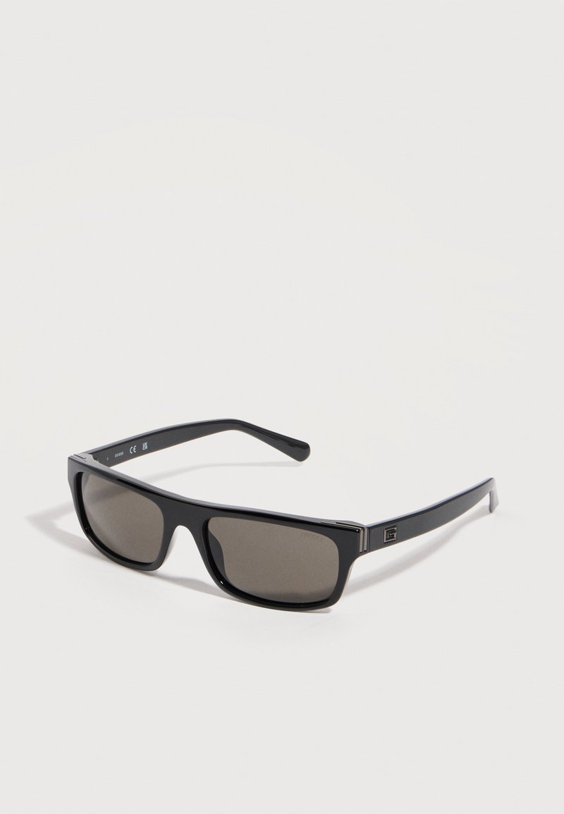 Lunettes de soleil rectangulaires noires avec des verres teintés foncés et une monture brillante, posées à plat sur une surface claire.