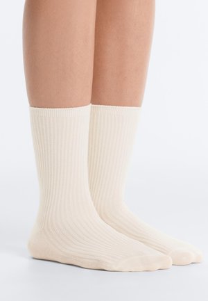 Lábak, középvádliig érő, bordázott krémszínű zoknit viselnek, egy fehér felületen állnak, egyszerű fehér háttér előtt.