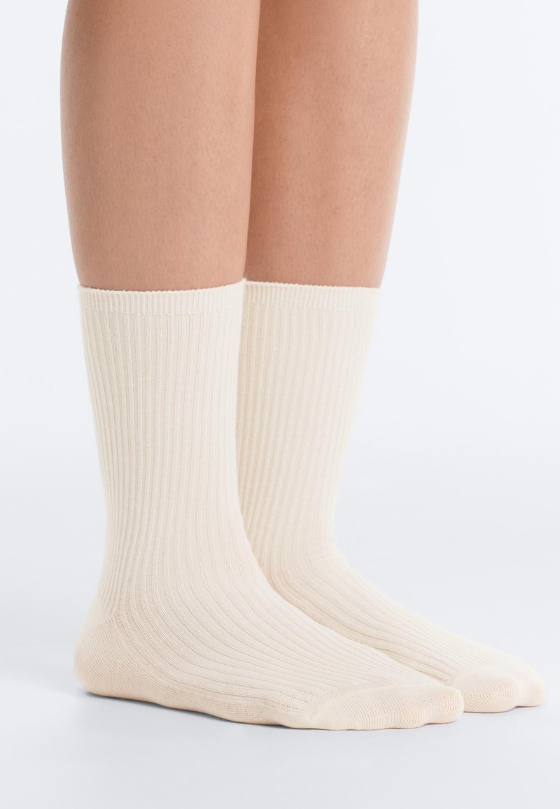 Jambes portant des chaussettes crème côtelées mi-mollet, debout sur une surface blanche avec un fond blanc uni.
