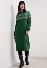 Dolga zelena pletena obleka s potiskom na yoku v kremasti, rjavi in modri barvi. Ima rebraste manšete in rob; v kombinaciji z črnimi škornji do kolen.